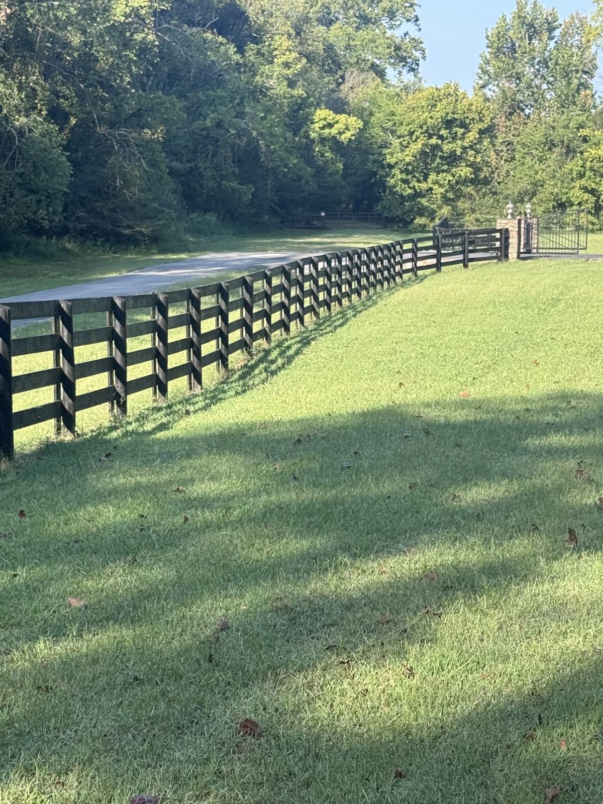Four-board wooden farm fence painted black, running along a driveway toward an iron entry gate